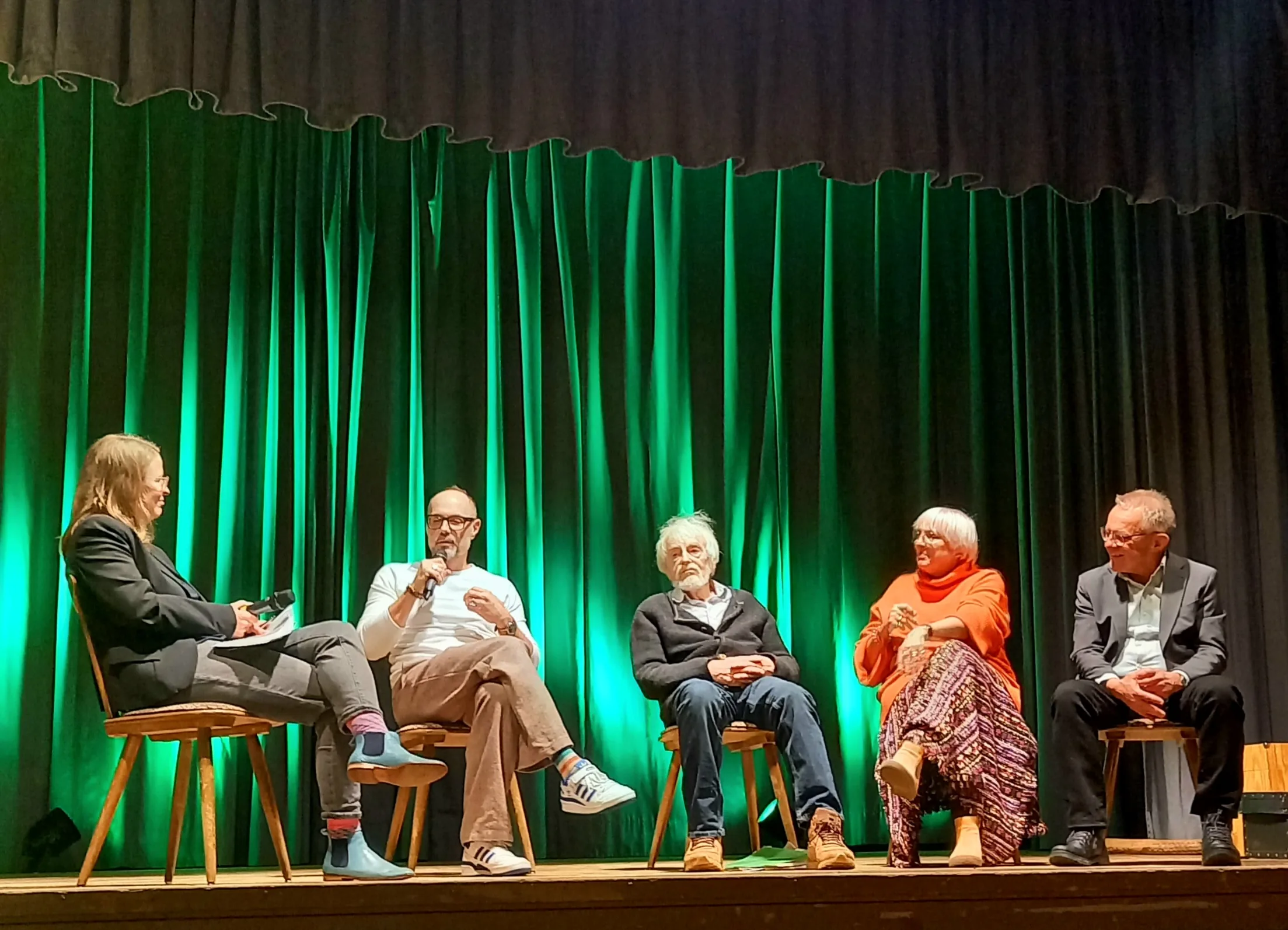 Podiumsdiskussion auf einer Bühne vor grünem Vorhang: Christina Mader, Volker Klüpfel, Michael Marmon, Claudia Roth und Thomas Gehring sitzen auf Holzhockern, Klüpfel spricht ins Mikrofon.