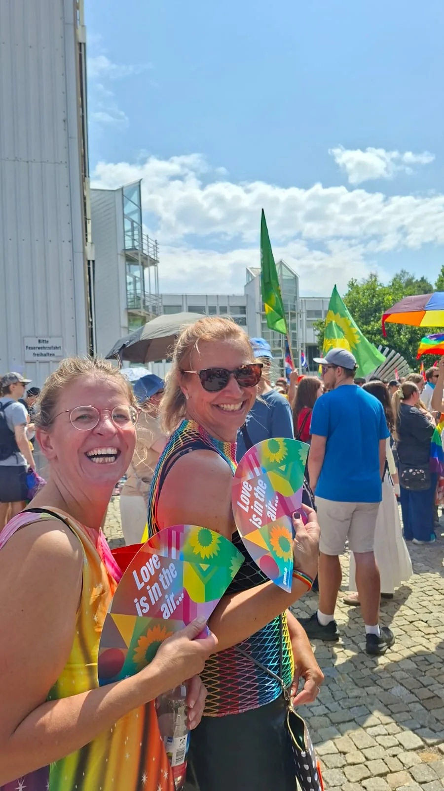 Zwei lachende Frauen im CSD-Zug halten runde Fächer mit Sonnenblume und der Aufschrift „Love is in the air…“ in die Kamera.