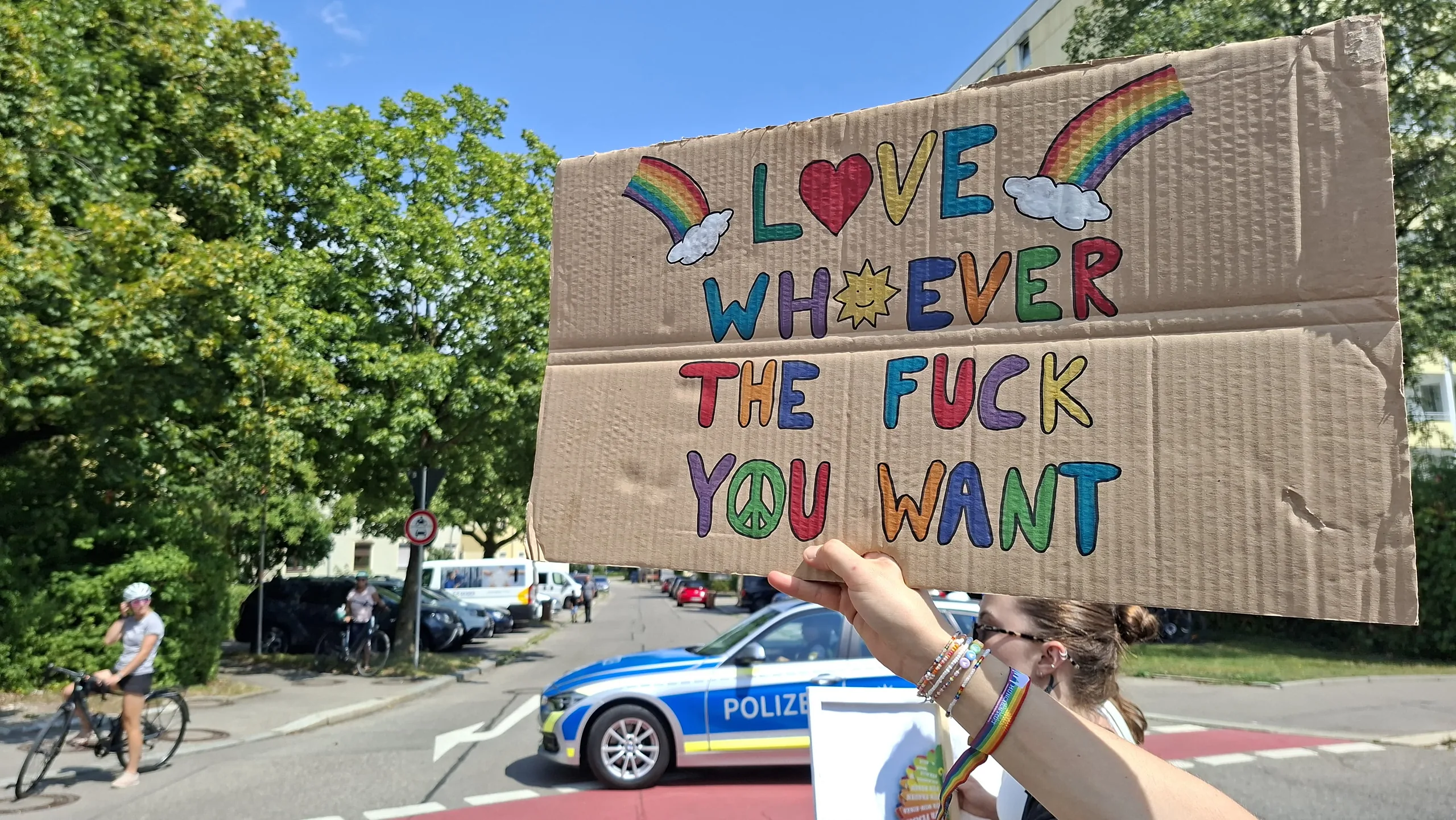 Handgemaltes Pappschild mit Regenbogen, Herz und Sonne und der Aufschrift „Love whoever the fuck you want“.