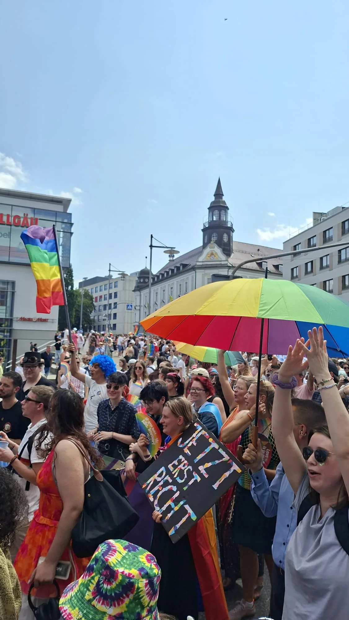 Dichte Menschenmenge beim CSD mit einem großen Regenschirm in Regenbogenfarben und einer Regenbogenfahne über den Köpfen.