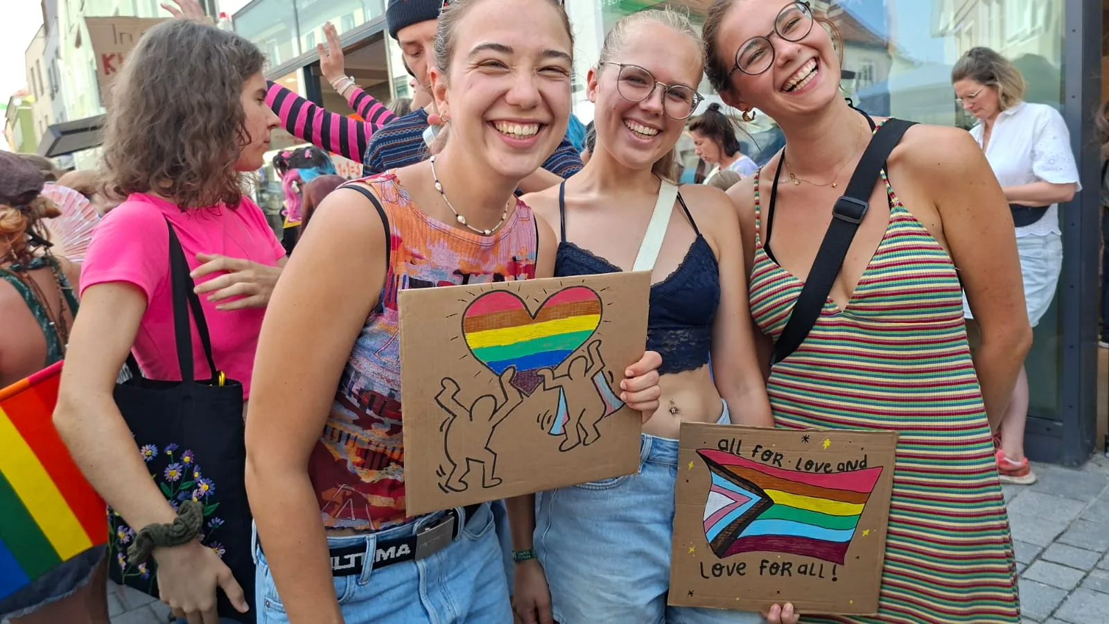 Drei lachende junge Frauen halten selbstgemalte Pappschilder: eines mit Regenbogenherz und tanzenden Figuren, eines mit Progress-Pride-Flagge und der Aufschrift „all for love and love for all!“.