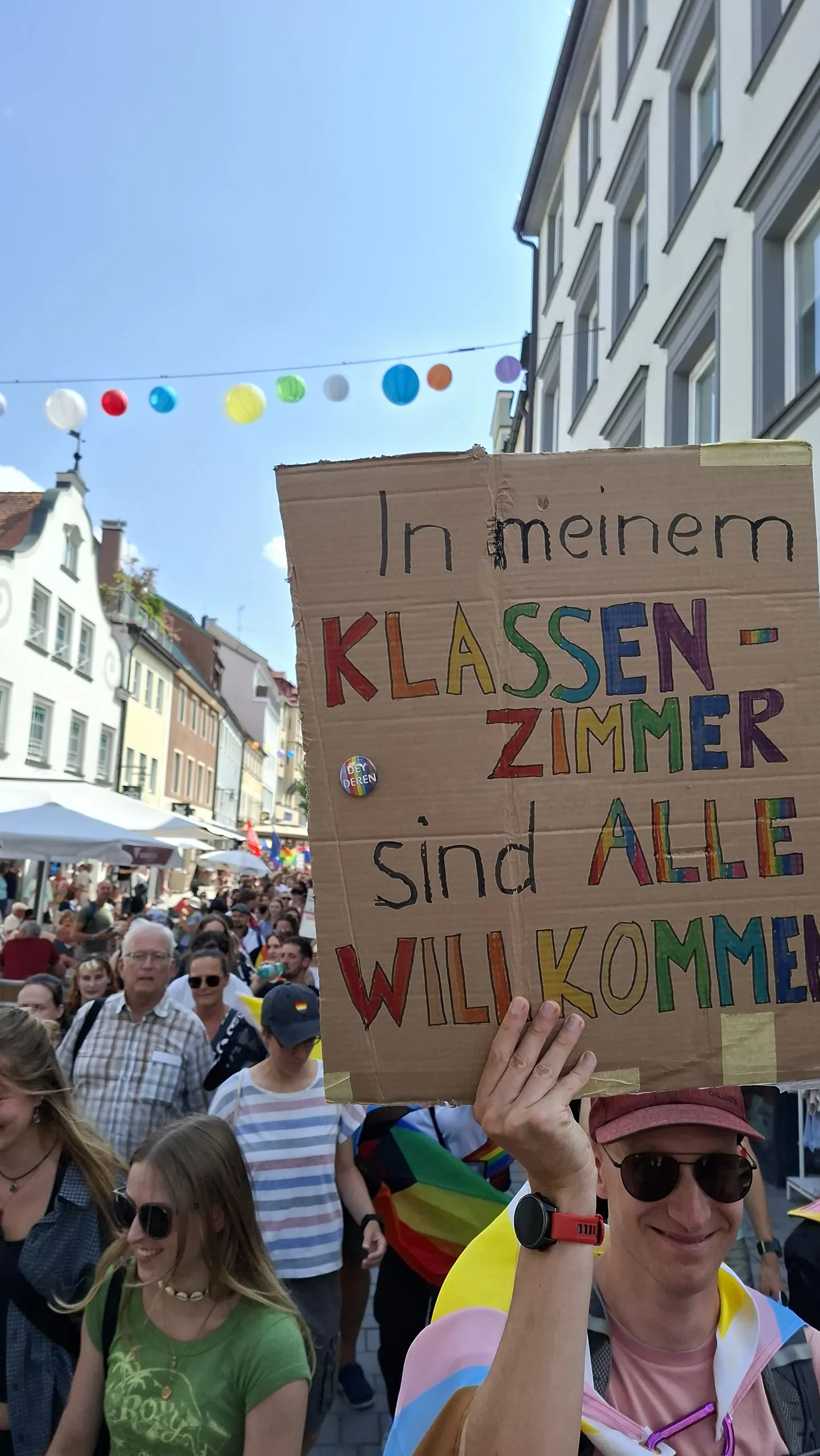 Über der Menschenmenge wird ein buntes Pappschild mit der Aufschrift „In meinem Klassenzimmer sind alle willkommen“ hochgehalten, dahinter Altstadthäuser.