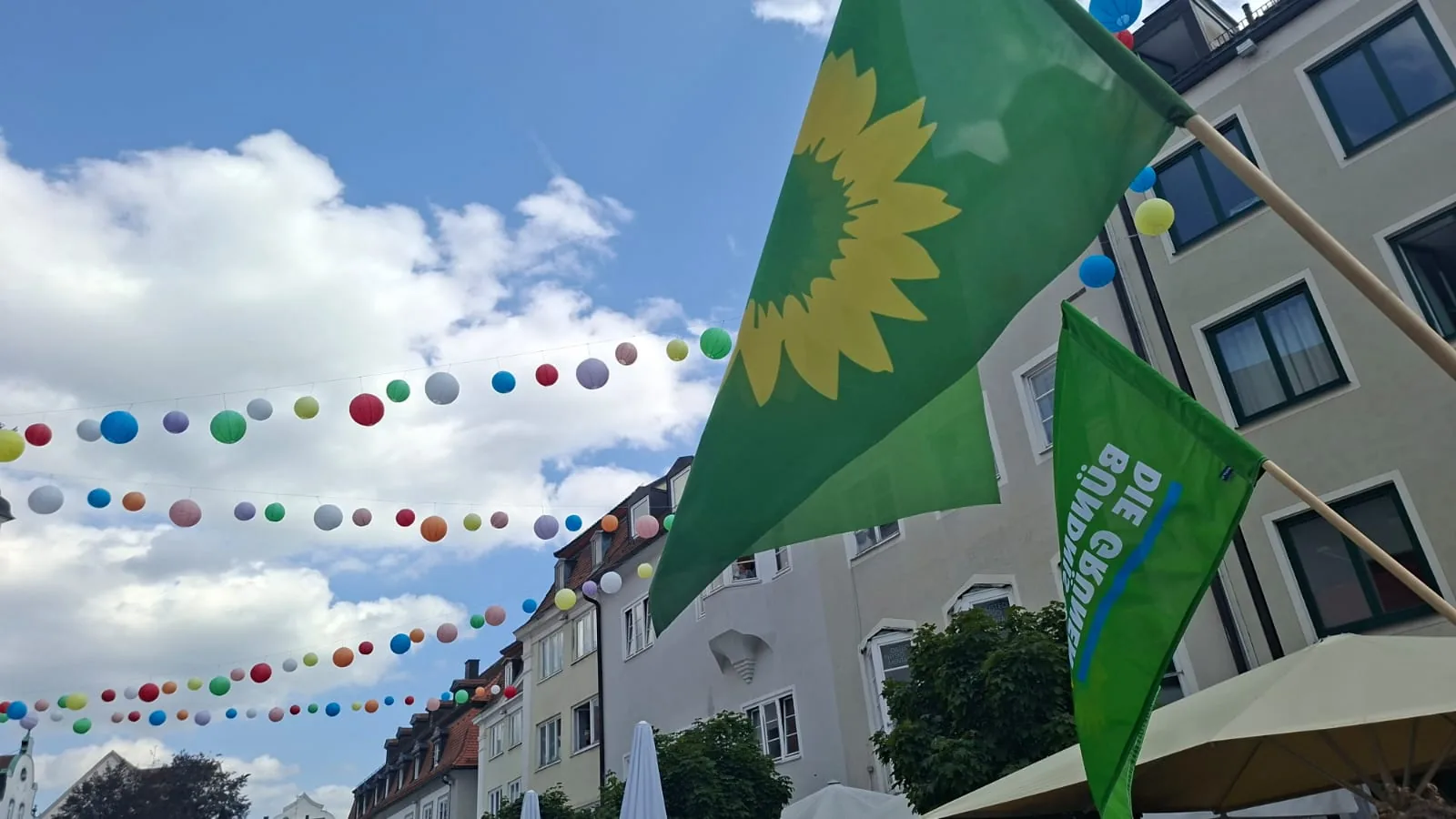 Grüne Fahnen mit der Sonnenblume von BÜNDNIS 90/DIE GRÜNEN wehen vor blauem Himmel, darüber bunte Ballongirlanden über der Straße.