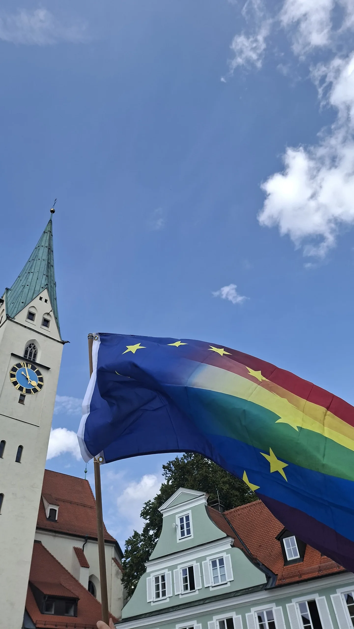 Eine Europafahne in Regenbogenfarben weht vor dem Turm der Kemptener Kirche mit der Turmuhr.