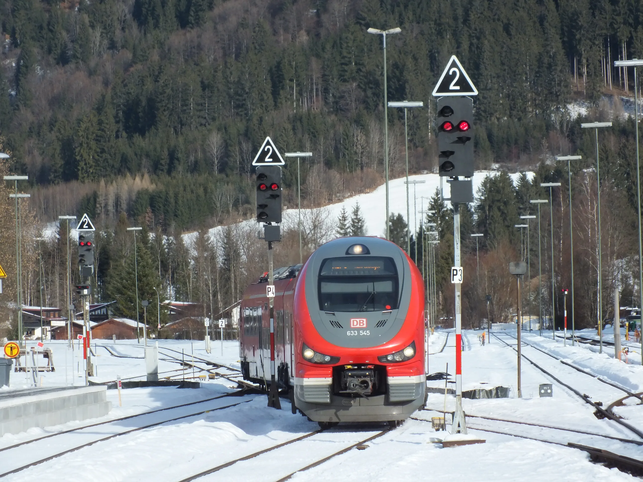 Ein roter Triebwagen der Deutschen Bahn steht im winterlichen Bahnhof Oberstdorf zwischen Signalen und schneebedeckten Gleisen, im Hintergrund Wald und Berghang.