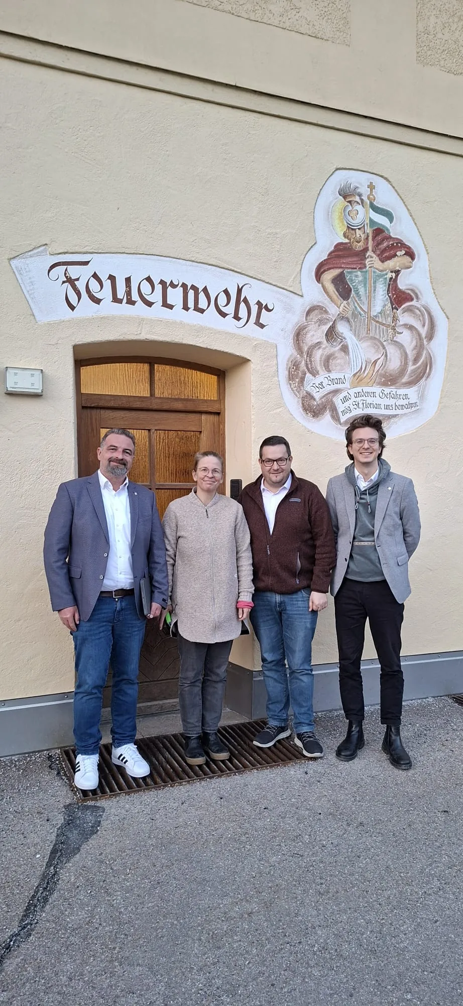Robert Gast, Christina Mader, Markus Adler und Leon Eckert stehen vor einem Feuerwehrhaus mit dem Schriftzug „Feuerwehr“ und einem Wandbild des heiligen Florian.
