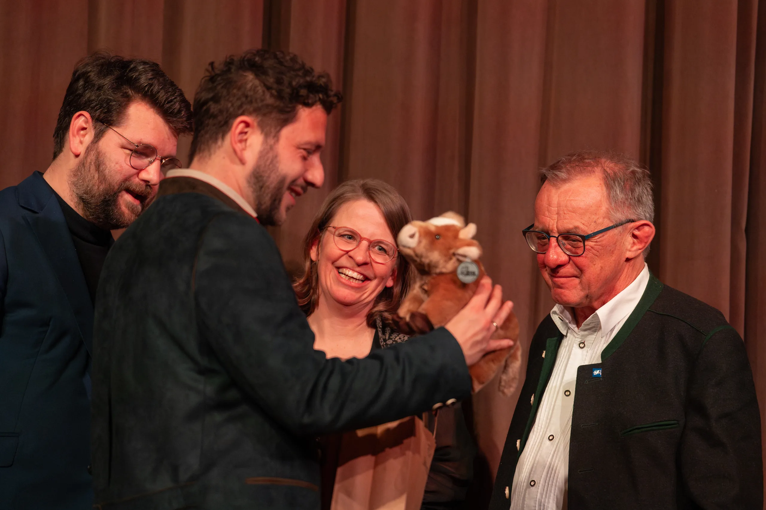 Vier Personen stehen lachend auf der Bühne, eine von ihnen hält eine Stoffkuh als Geschenk in den Händen.