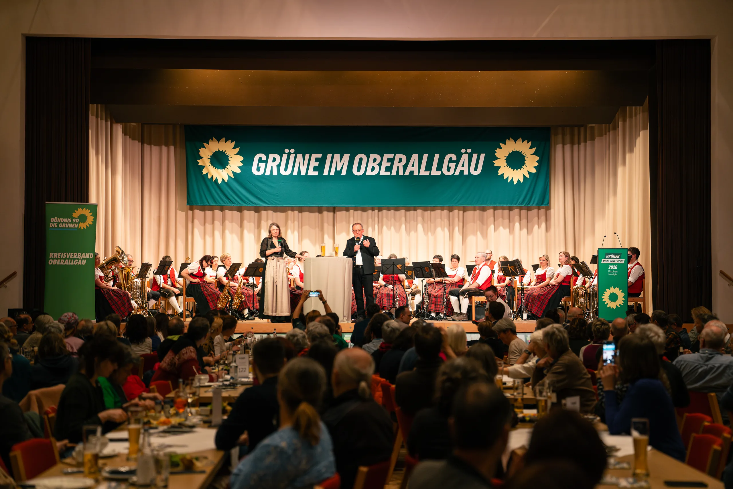 Blick von hinten in den vollen Saal auf die Bühne mit dem Transparent „GRÜNE IM OBERALLGÄU“ und der Blaskapelle in roter Tracht.