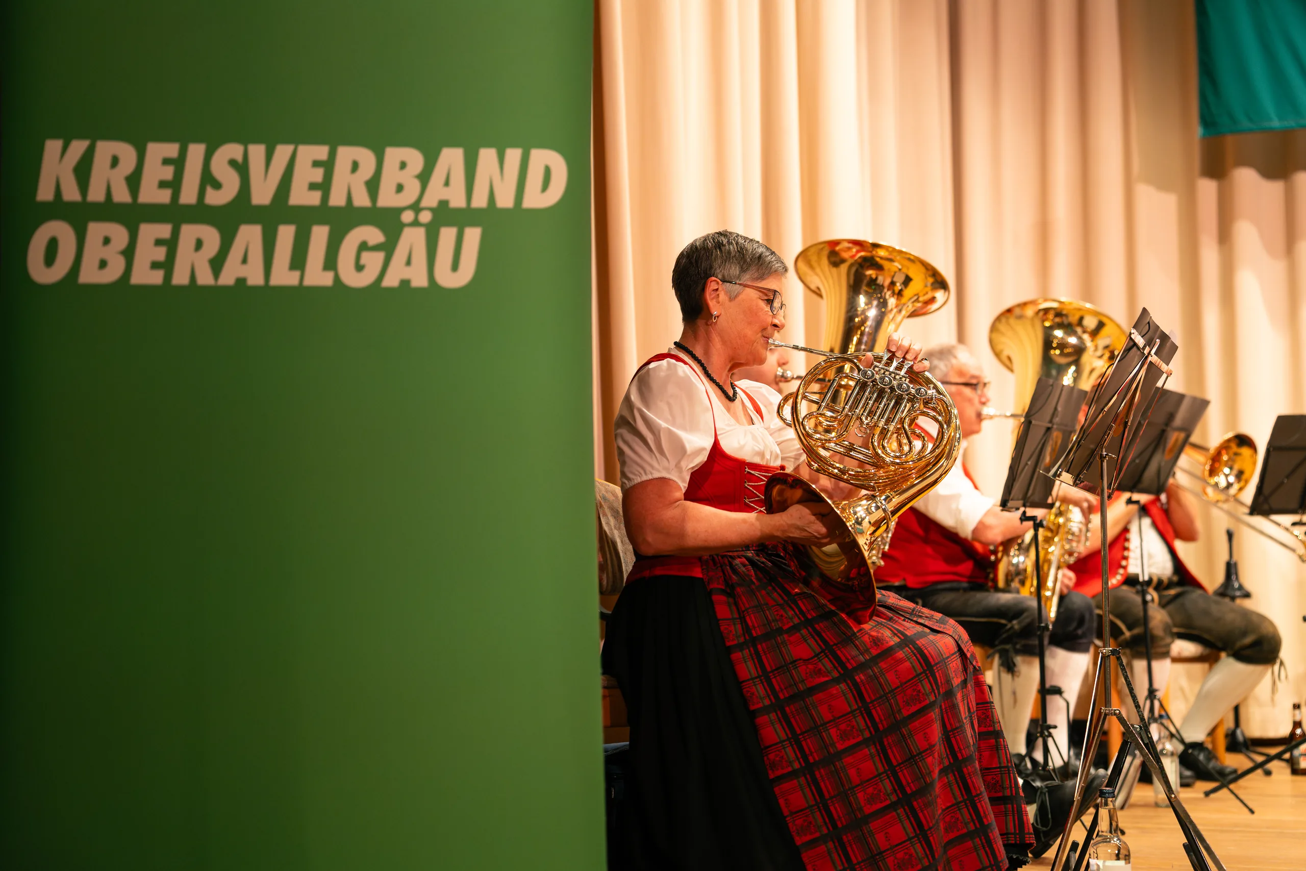 Eine Musikerin in roter Tracht spielt Waldhorn neben einem grünen Aufsteller mit der Aufschrift „Kreisverband Oberallgäu“.