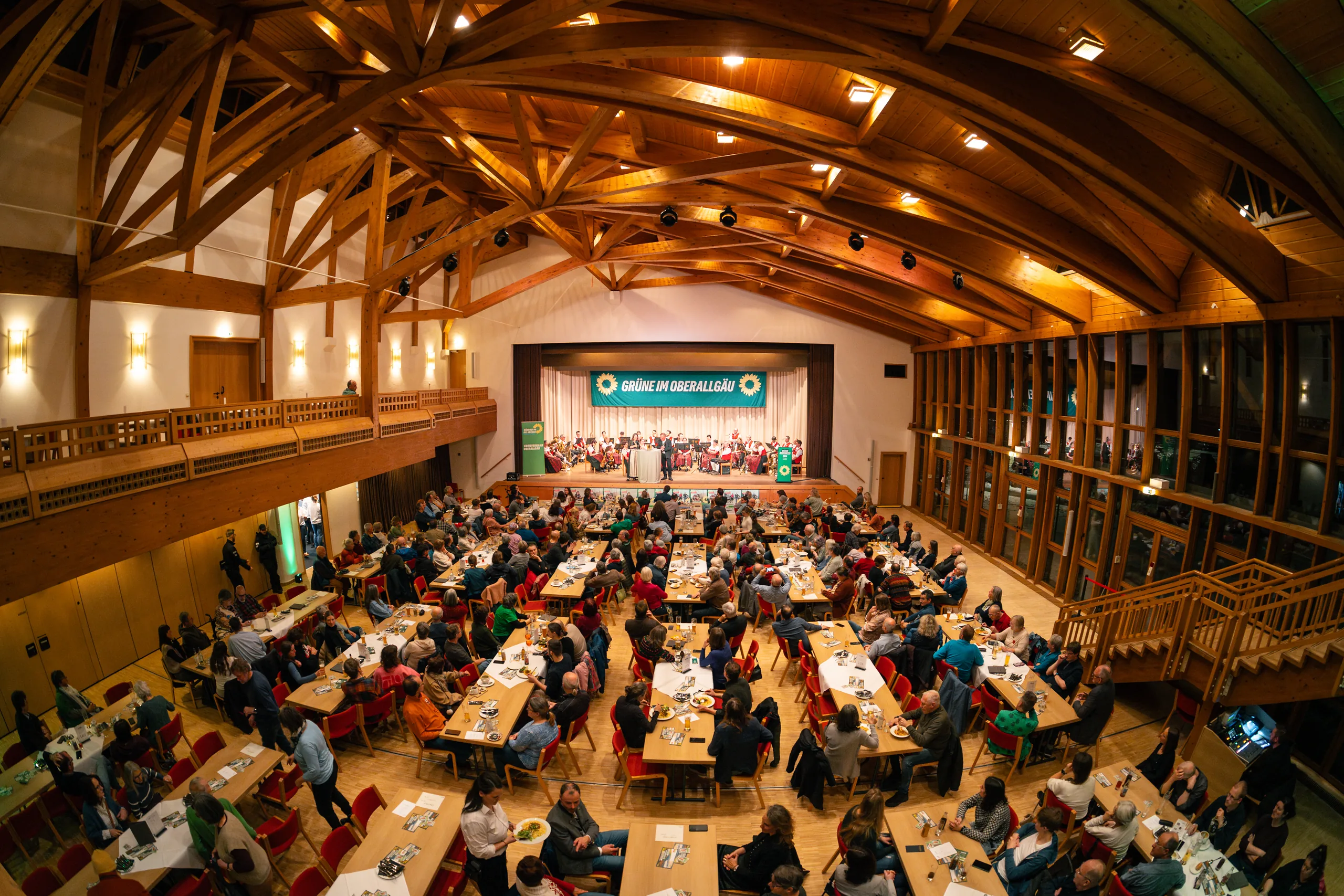 Blick von der Empore in den vollbesetzten Saal mit hölzerner Bogendecke; an langen Tischen sitzen die Gäste, vorn die beleuchtete Bühne.