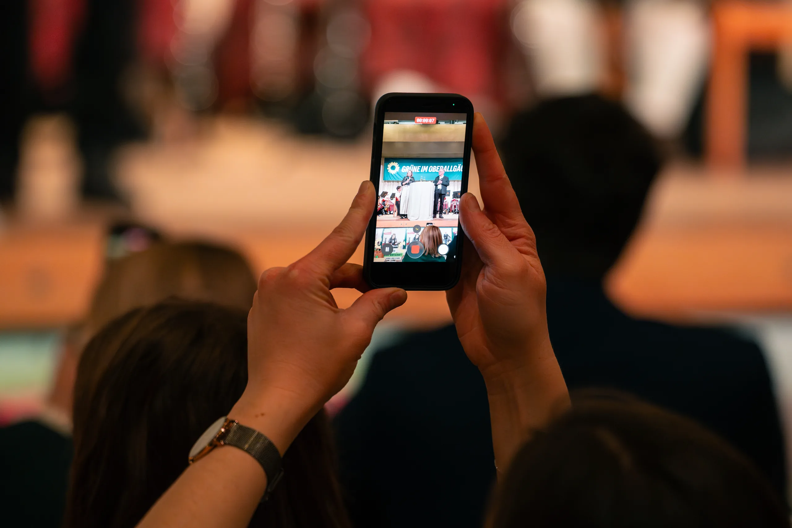 Zwei Hände halten ein Smartphone hoch und filmen die Bühne, im Vordergrund unscharf die Köpfe zweier Gäste.