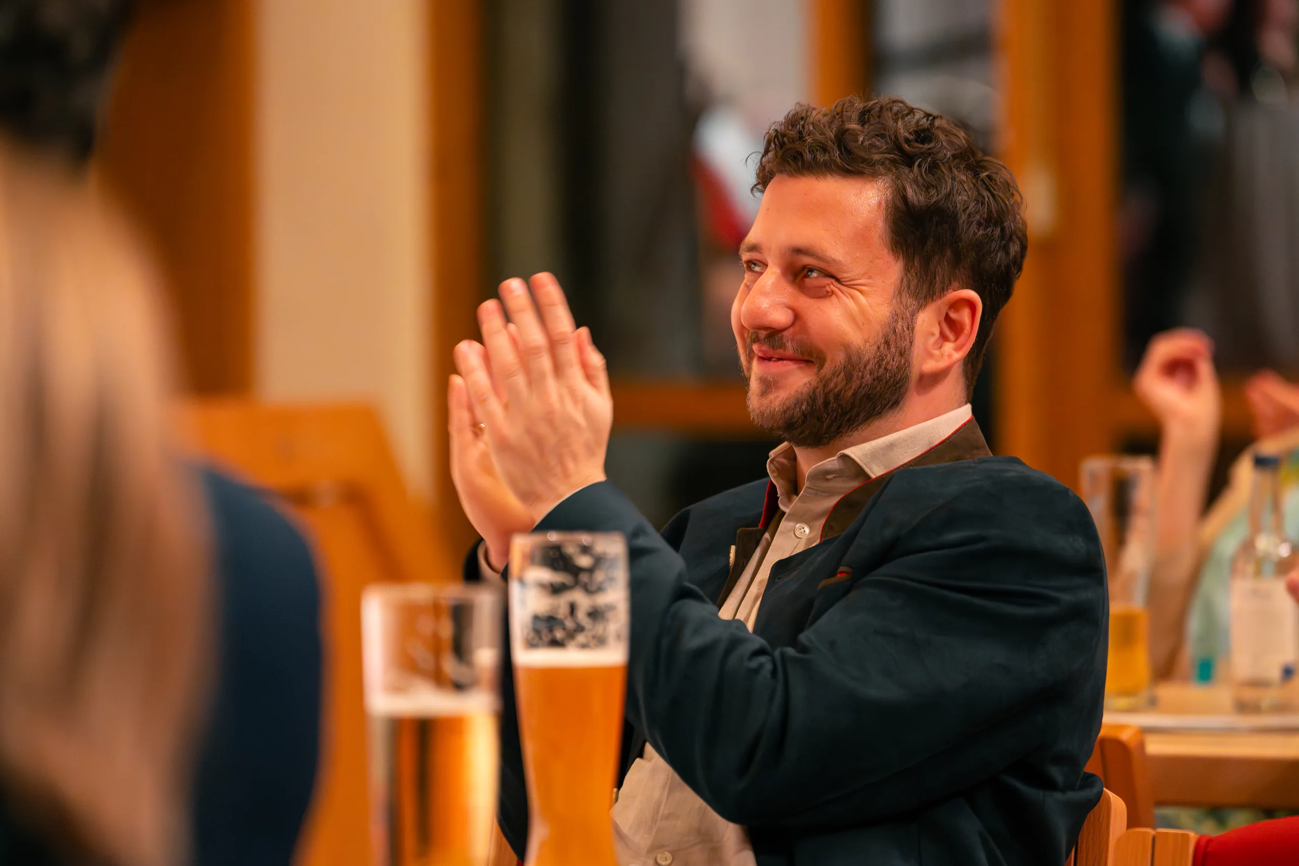 Ein Gast im dunklen Sakko applaudiert lächelnd an einem Tisch mit Weißbiergläsern.