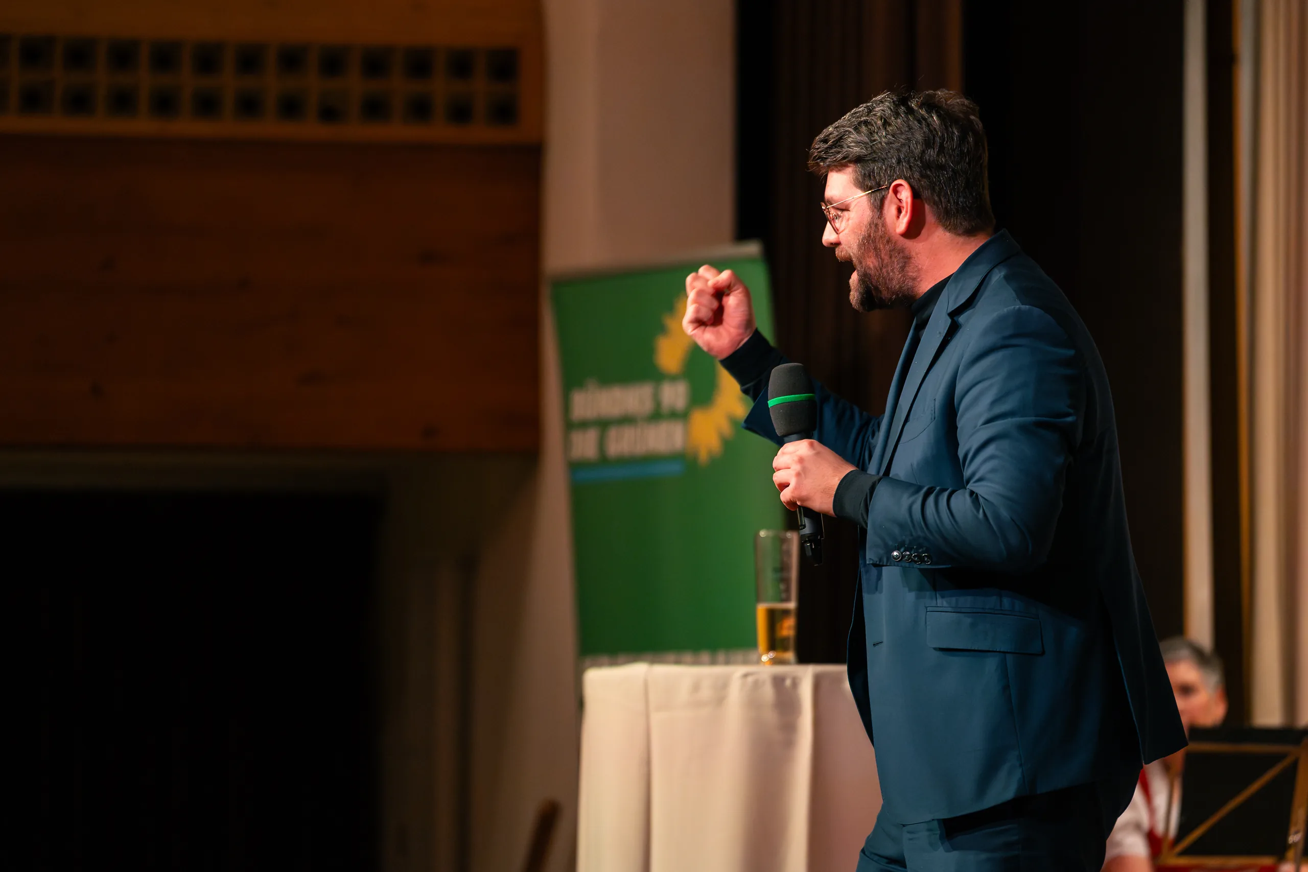 Ein Redner spricht mit erhobener Faust ins Mikrofon, hinter ihm ein grüner Aufsteller mit Sonnenblume.