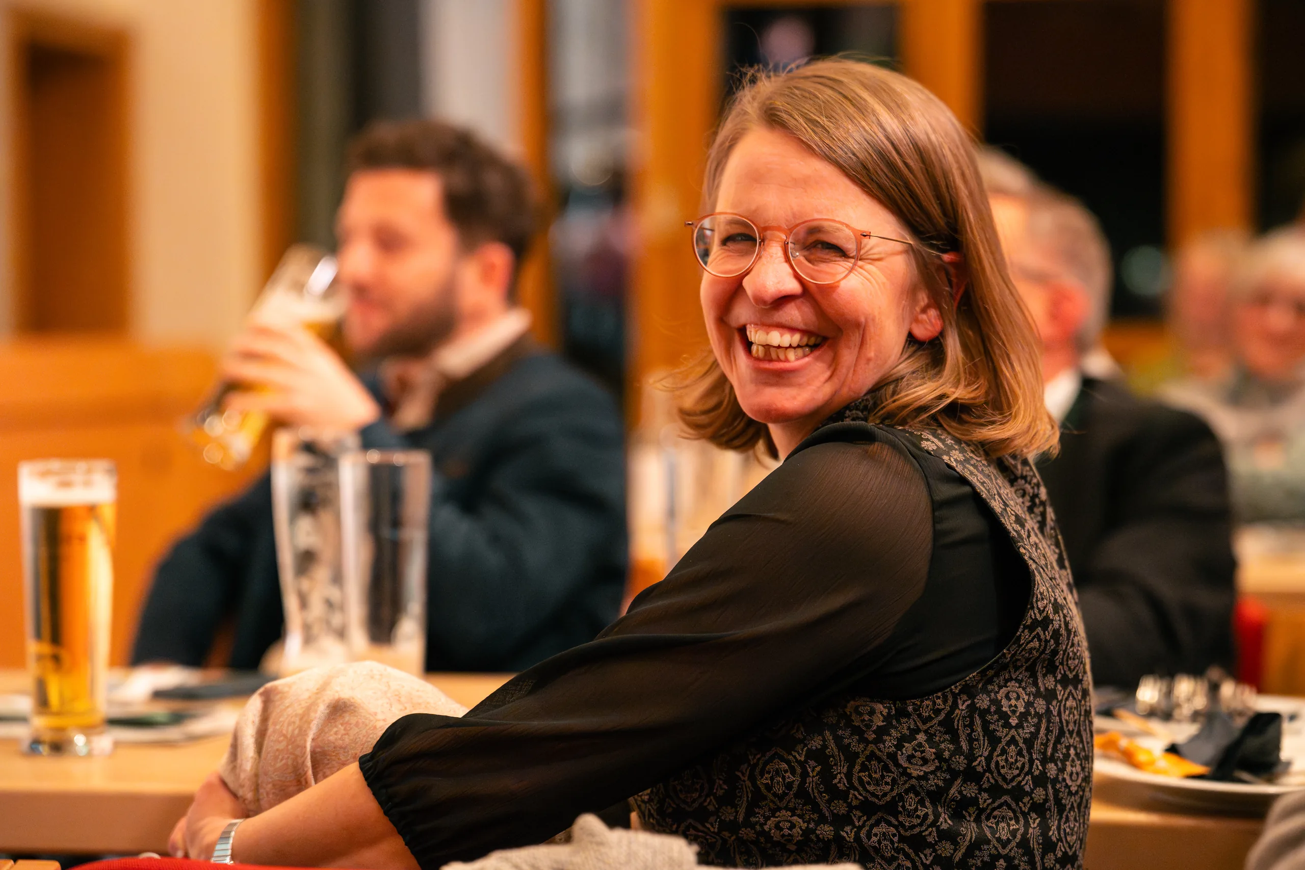 Eine Besucherin lacht an einem Tisch mit Weißbiergläsern in die Kamera, dahinter weitere Gäste.